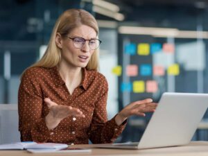 Besorgte Geschäftsfrau mit Brille arbeitet am Laptop im modernen Büro und reagiert frustriert auf einen Computerfehler oder Systemabsturz. Bürohintergrund mit Notizzetteln an der Glaswand, Problem im Arbeitsalltag, digitale Herausforderungen am Arbeitsplatz.