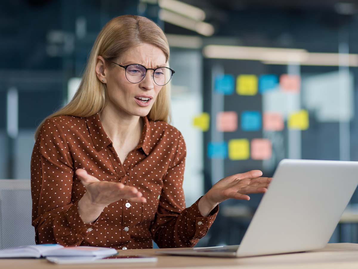 Besorgte Geschäftsfrau mit Brille arbeitet am Laptop im modernen Büro und reagiert frustriert auf einen Computerfehler oder Systemabsturz. Bürohintergrund mit Notizzetteln an der Glaswand, Problem im Arbeitsalltag, digitale Herausforderungen am Arbeitsplatz.