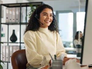 Lächelnde IT-Mitarbeiterin mit Headset am Arbeitsplatz bei der Fernwartung in Unternehmen über Remote Desktop.