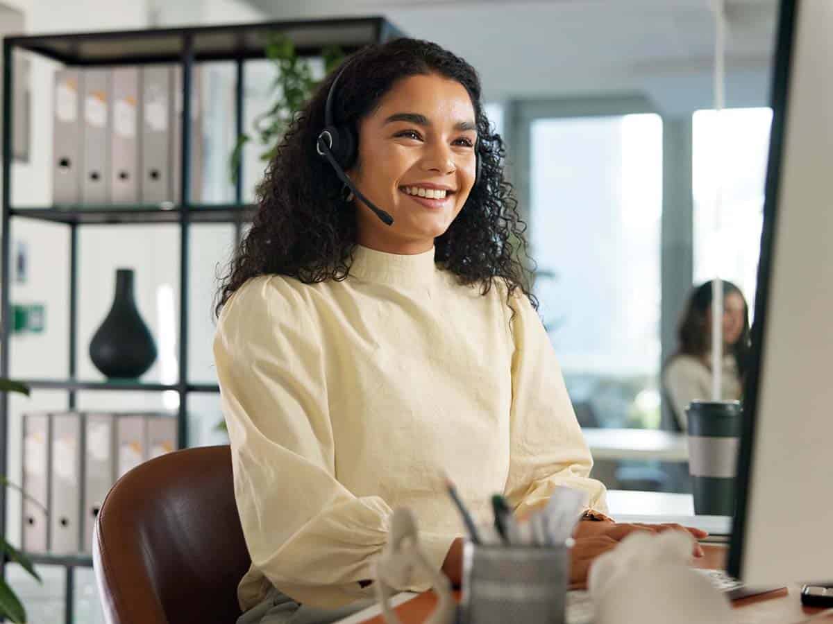 Lächelnde IT-Mitarbeiterin mit Headset am Arbeitsplatz bei der Fernwartung in Unternehmen über Remote Desktop.