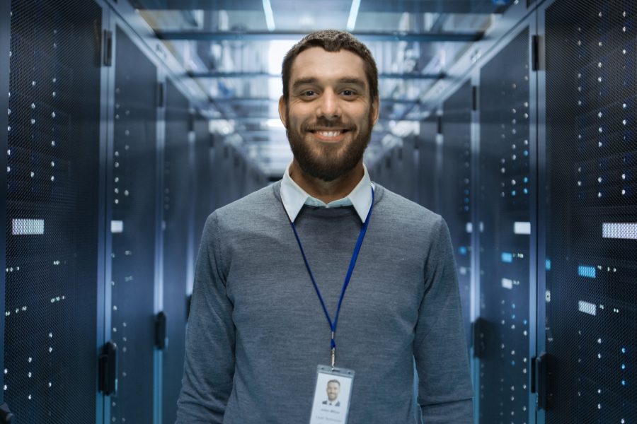 Portrait of a Curios, Positive and Smiling IT Engineer Standing in the Middle of a Large Data Center Server Room.